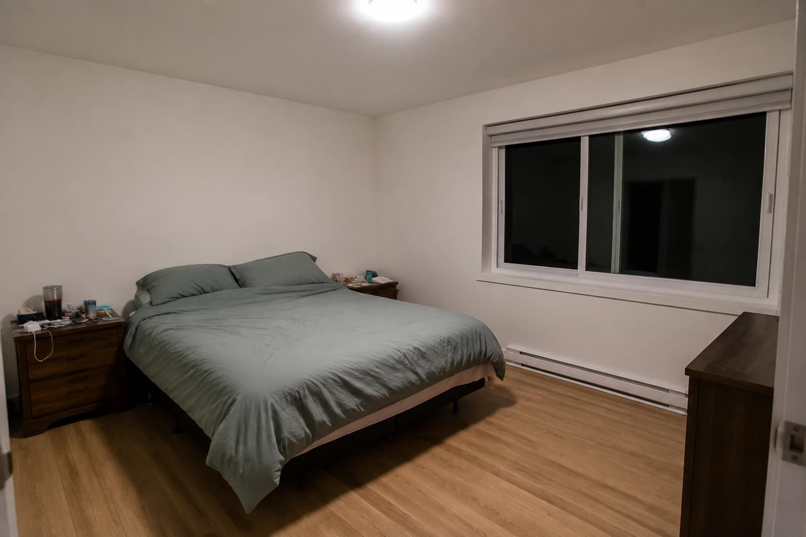 A bedroom lit only by a single bright ceiling fixture, with cluttered nightstands and no rug