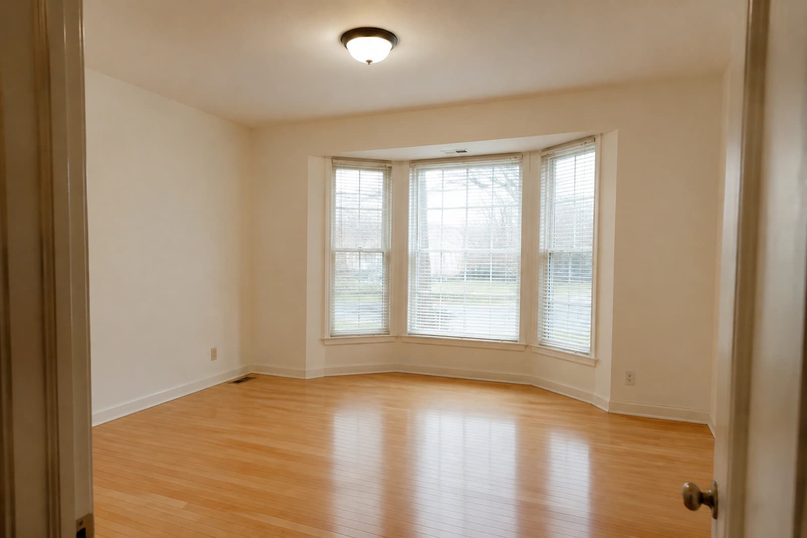 An empty living room with white walls, a bay window and pale hardwood floors, photographed on a phone in flat daylight